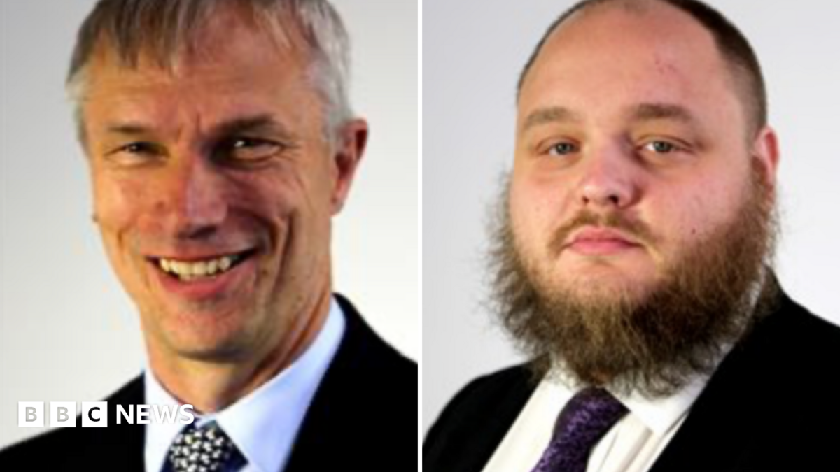 A side by side shot of two men in suits. The man on the left has short grey hair and the man on the right has a skin fade with a short unruly black beard