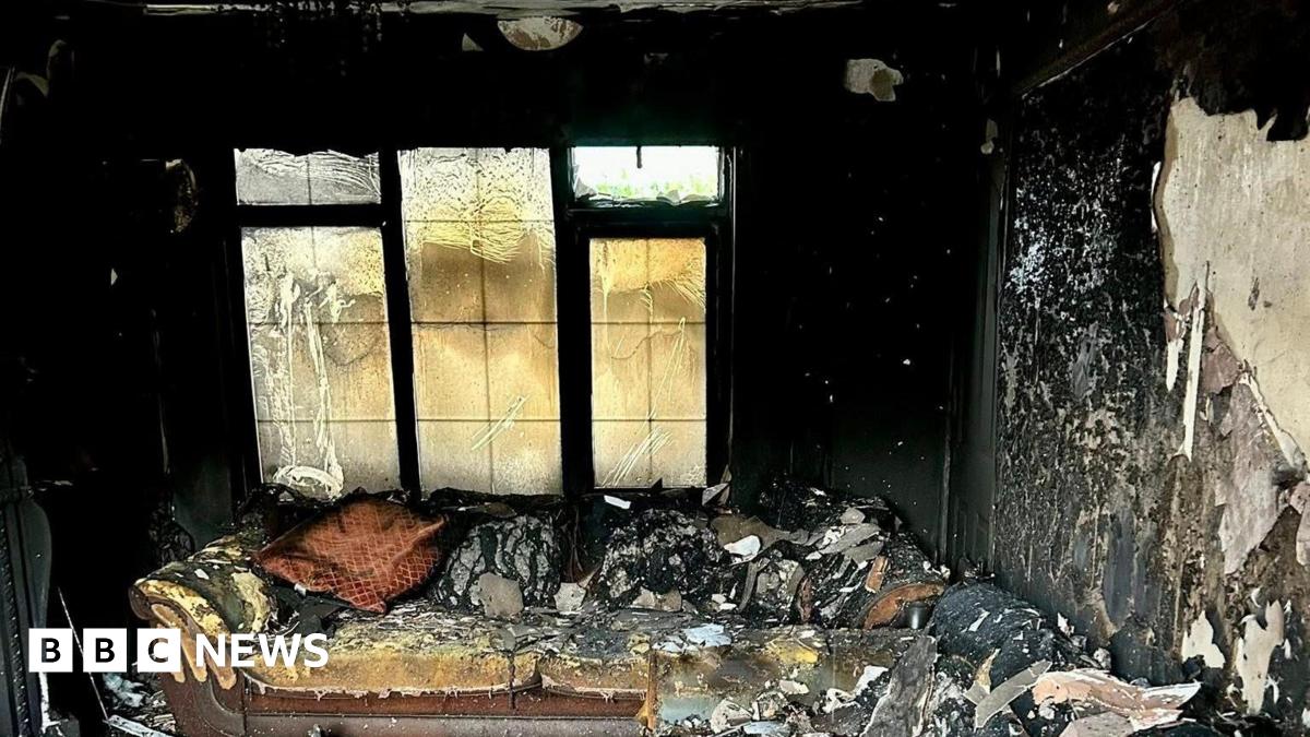 The inside of a living room which has been ravaged by fire. Huge chunks of paint and plaster are falling off the walls, the sofa is singed and the window is warped. 