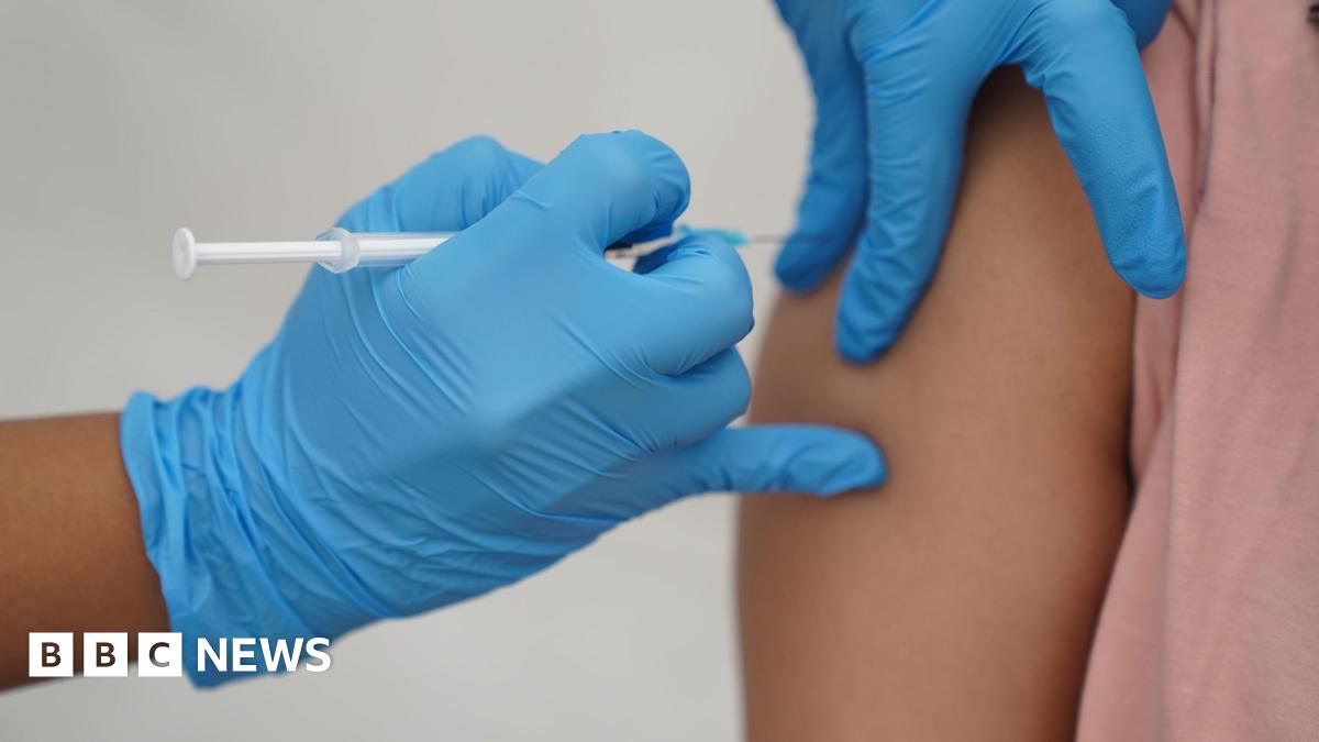 Close-up of a vaccine being administered. The healthcare worker wears blue gloves and the recipient is wearing a pink top.