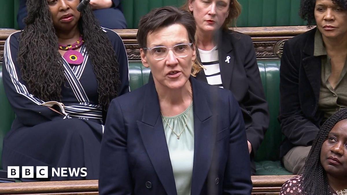 A woman is stood in the Houses of Parliament. She is wearing glasses and has short dark hair. She has a blue blazer on with a mint green shirt underneath 