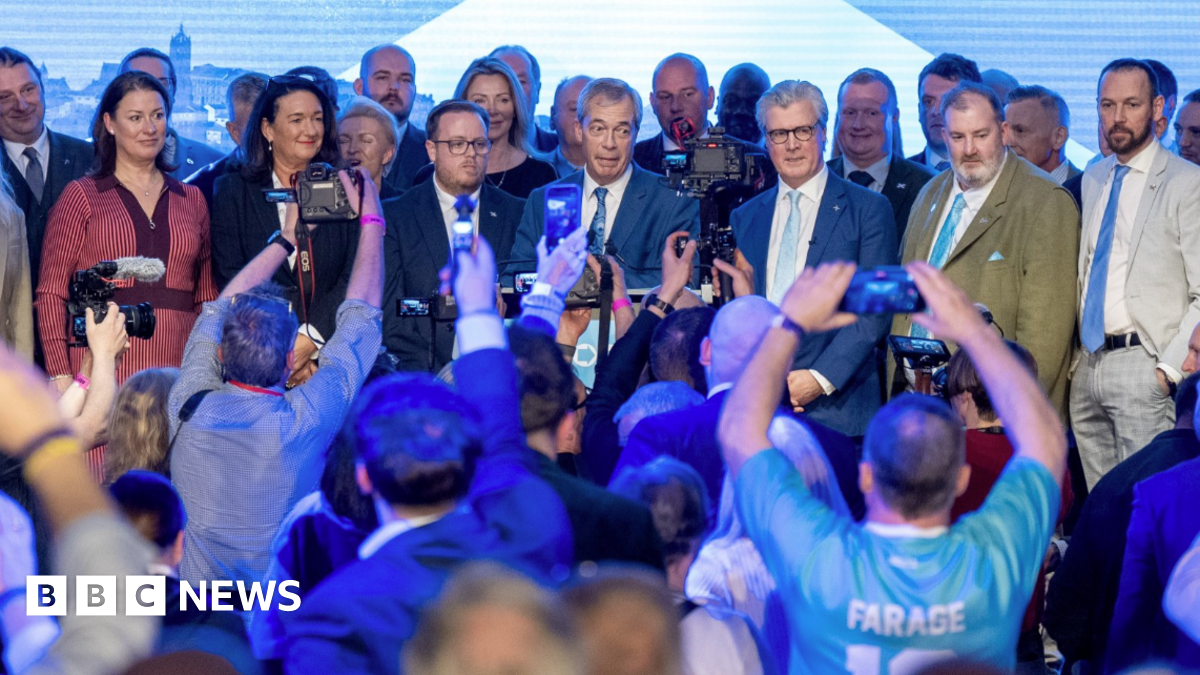 A large crowd with many attendees holding up phones and cameras to photograph a group of people standing on a stage beneath bright lighting and a large screen backdrop