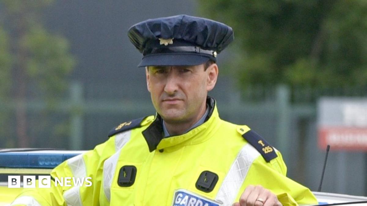 A man wearing an Irish police officer uniform of a yellow hi-vis jacket standing behind the open door of a police car which is stationary.