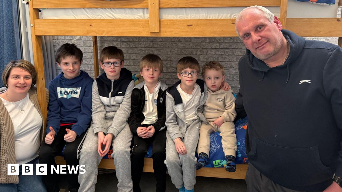 Mother Sam stands to the far left wearing a white T-shirt, cardigan and jeans. She has a short bob hairstyle. Next to her, sitting on the bottom bunk bed are her five sons all lined up together wearing tracksuits. Their father Jason is standing on the...