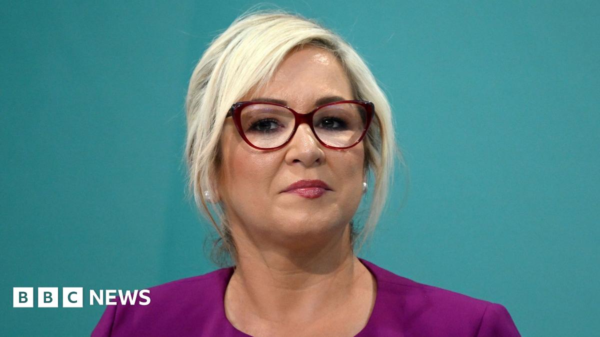 Michelle O'Neill is standing in front of a green backdrop. She's wearing red glasses and a pink jacket with pearl earrings. 