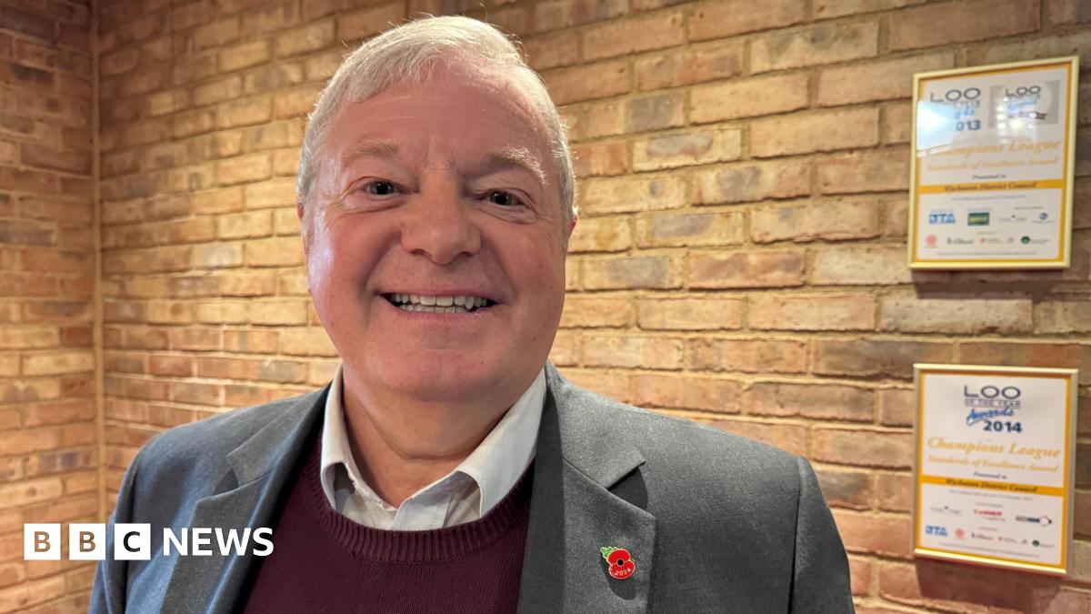 Councillor Alan Amos is smiling at the camera, wearing a grey suit over a maroon jumper. He has short, neatly brushed white hair.