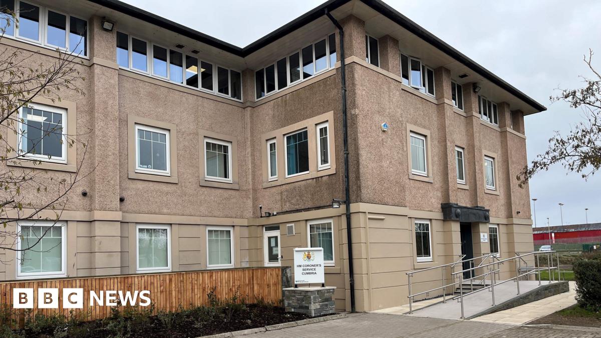 The entrance to Cumbria coroner's court building. The entrance is at the left-hand side of a three-storey building, with beige cladding on the ground floor and shelldash on the upper floors. There is a sign for the coroner's court in front on a ramp le...