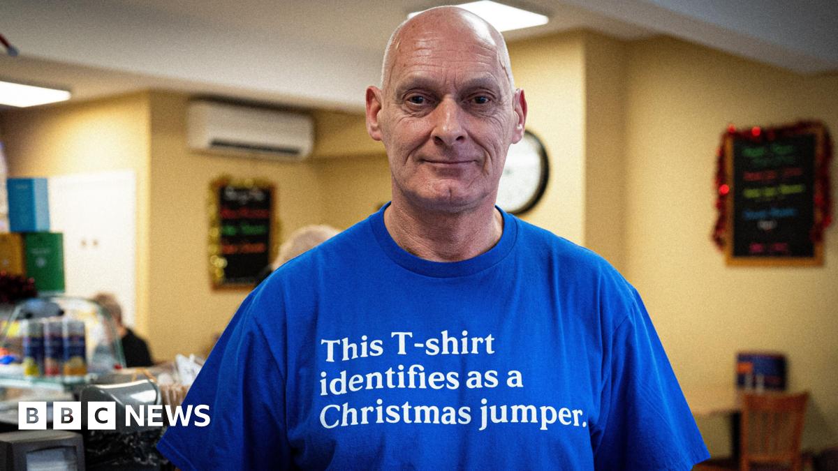 A bald man in a royal blue T-shirt bearing the words "This T-shirt identifies as a Christmas jumper" stands with his back to the counter in his coffee shop, a customer  on the other side of it. 