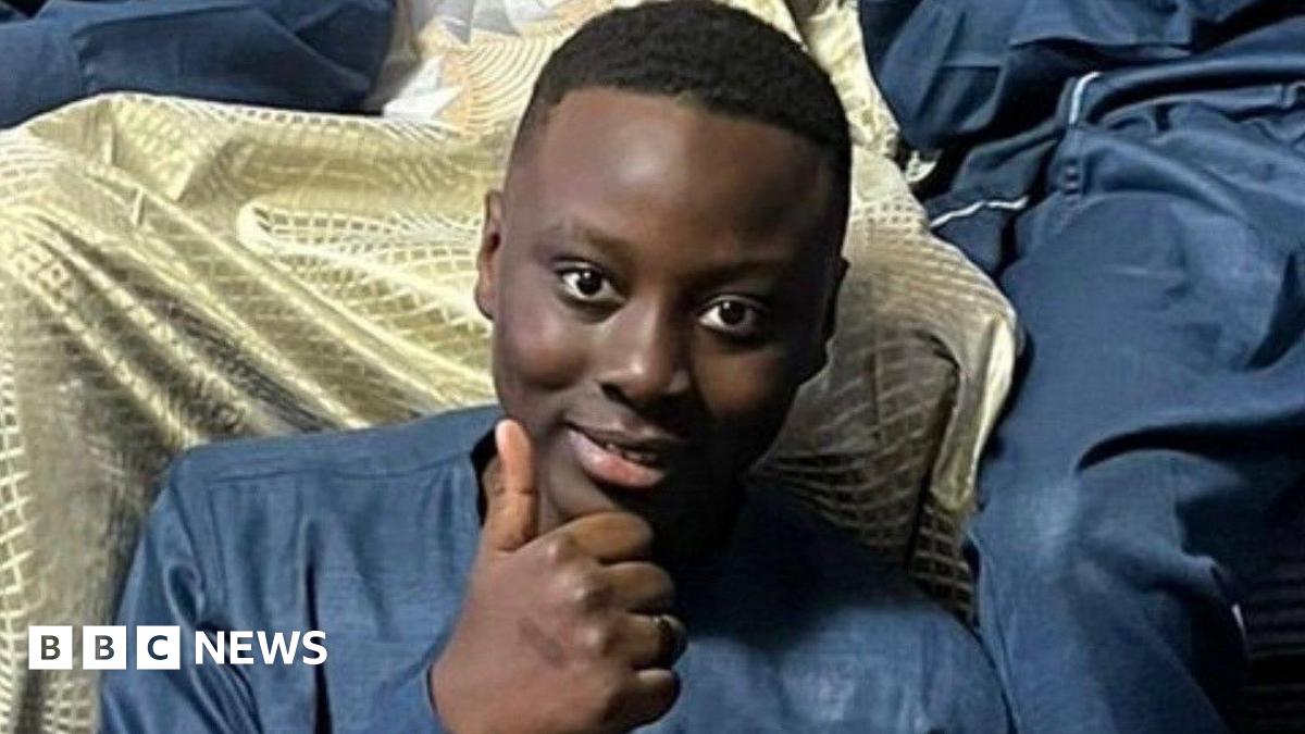 A young teenager sits smiling and giving a thumbs up to the camera. 