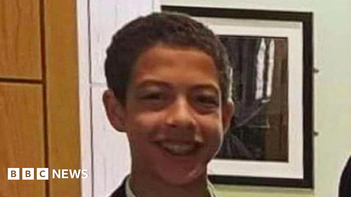 Noah is smiling in front of a green wall with a picture behind him that is reflecting a window. He has brown short hair and brown eyes. 