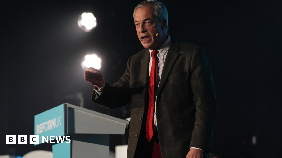 Nigel Farage wearing a brown suit and a red tie stands at a Reform UK podium.