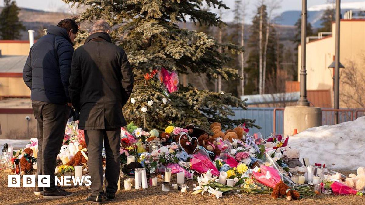 Two men standing outside, backs to camera looking down at a large collection of flowers and presents left for the victims of the school shooting, placed underneath a large tree.