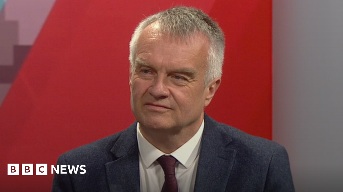 A man with short grey hair stands on a pavement with parked cars on the road. He is wearing an open-collared shirt under a brown jacket.