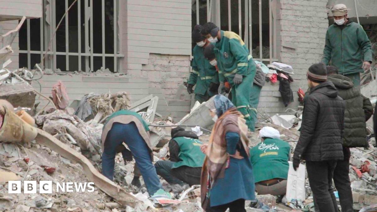 About six people in green jackets look through the rubble outside a building. The remains of two windows with vertical white bars across the holes can be seen. Another three men are looking on and a woman walks past. 