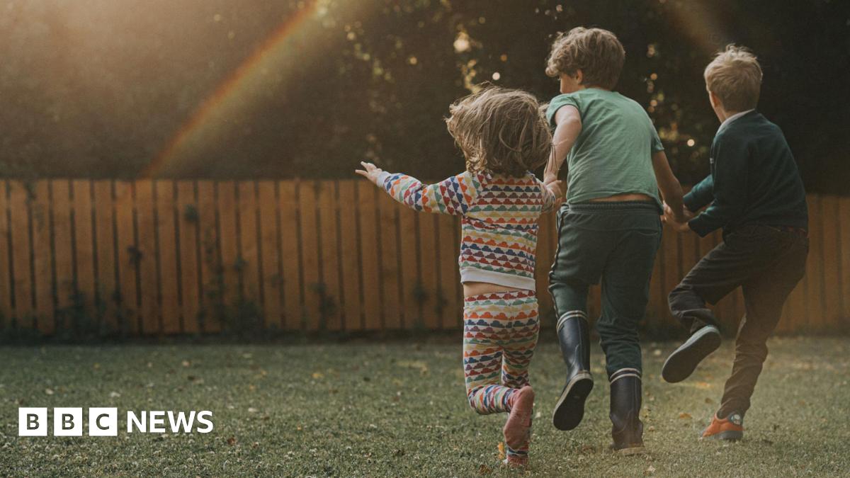Three children in a garden run towards a fence, with the picture taken behind them. The sun creates a rainbow-like effect.