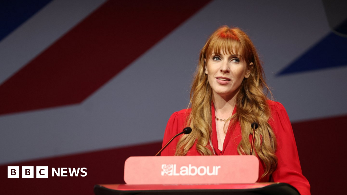 Angela Rayner addresses the Labour Women's Conference in Liverpool last year. She stands at a podium with Labour's red rose logo, wearing a bright red dress, in front of a Union Jack flag. She wears her long hair loose and styled into curls.