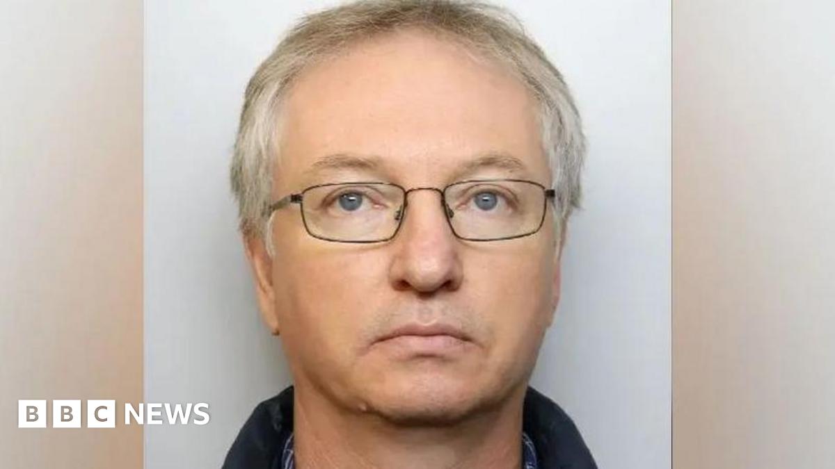 A custody picture of Chapman. He has no expression on his face, and wears dark, thin-framed glasses. 