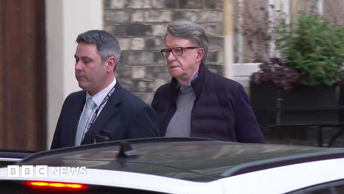 Lord Mandelson being arrested at his London home on Monday - He has dark grey hair swept back, is wearing glasses,  and has a serious look on his face as he walks away from his house