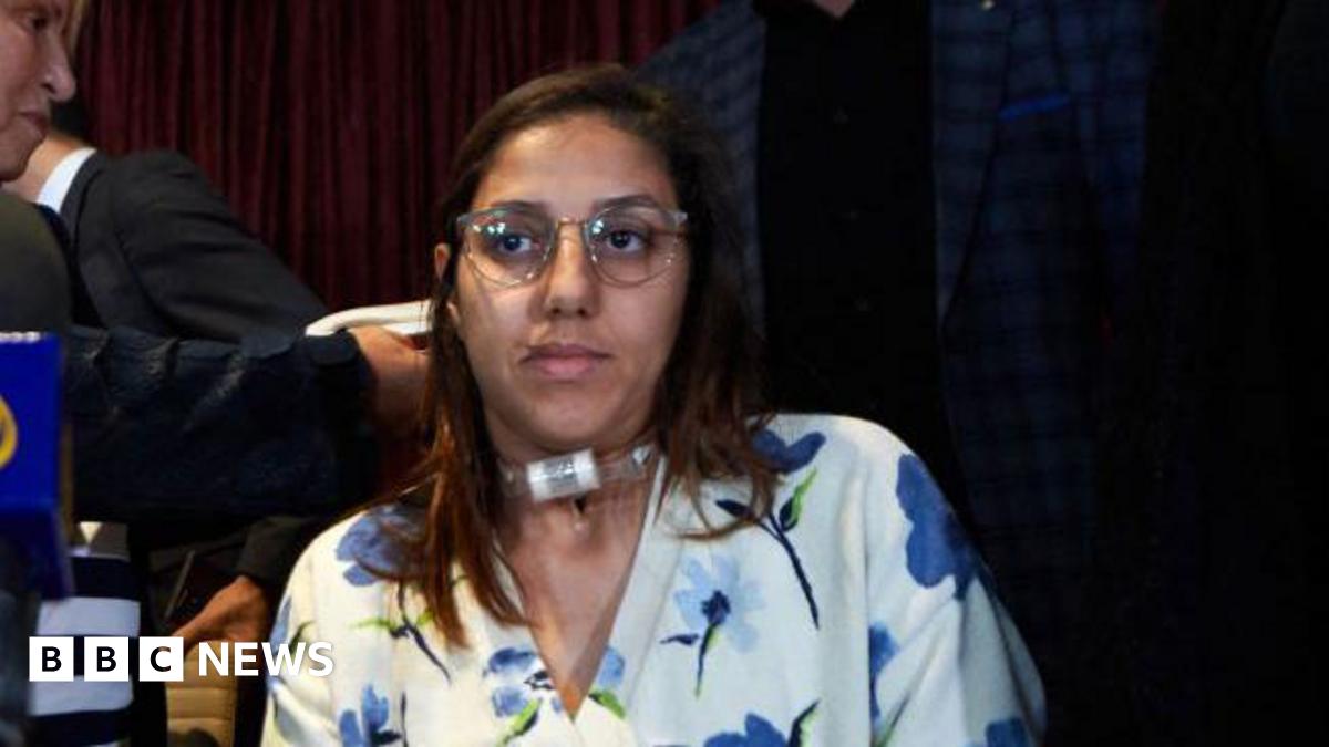 Saada Arbane attended a press conference with her lawyer (face partially seen in top left of image). Ms Arbane is visible from chest up; she has a visible speaking tube on her throat, long brown hair, spectacles and is wearing a white blouse with blue...