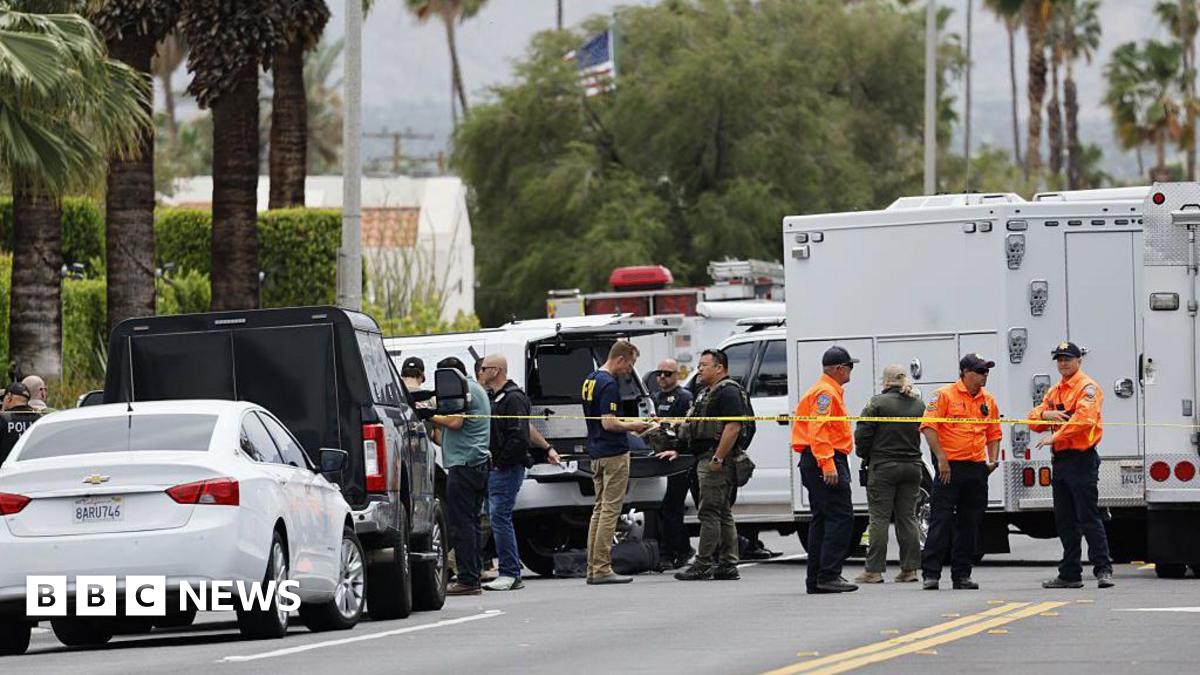 First responders in FBI shirts and orange uniforms stand on a palm tree-lined street. There are many trucks and a section of the stree is blocked off with yellow caution tape.