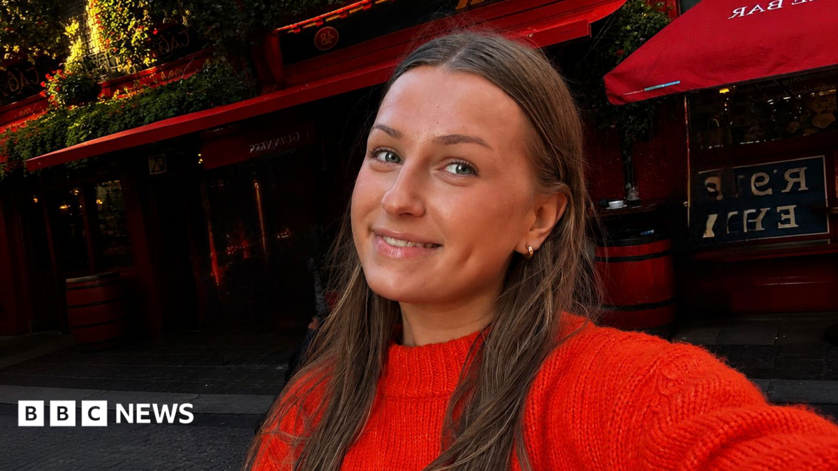 A selfie of Kelis Bailey who has long brown hair and is wearing a red jumper while standing in the street. She is holding the camera and taking the photo herself. 