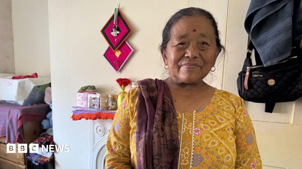 A Gurkha widow is wearing a bright yellow dress with a red shawl draped over her right shoulder. She also has a gold stud in her left nostril. Staring straight into the camera she's sitting in her bedroom and you can see the vague outlines of a few glass jars over her shoulder as well. A beautiful deep red shawl thrown over her right shoulder Indumati Thapa is standing in her small bedroom. Wearing a yellow dress you can see the foot stool she sits on when not in her bed behind her.