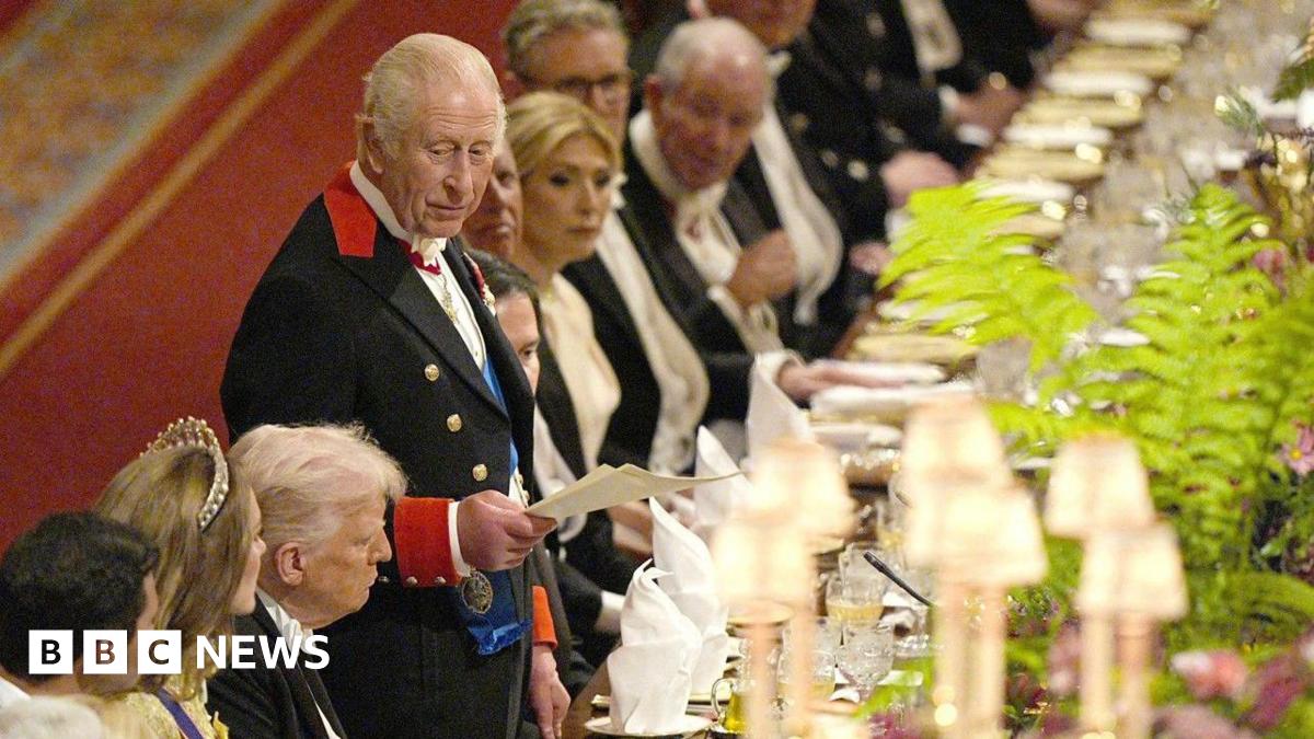 King Charles stands at a large banquet table and appears to be delivering a speech from a piece of paper. The US president Donald Trump is sat in a chair next to him. 