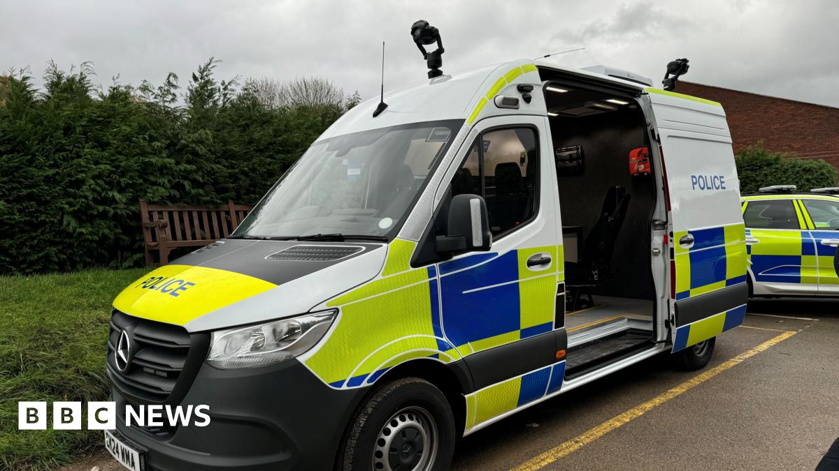 A van with blue, yellow and white police markings. It's middle door is open and two cameras are mounted to the top of it. It is parked next two another police car and a grass verge.