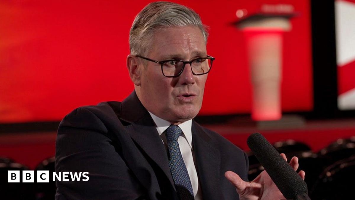 Sir Keir Starmer, dressed in a dark suit, white shirt, and blue patterned tie, is seated and speaking into a microphone during an interview at the Labour Party conference. Behind him is a red backdrop with blurred elements of the Union Jack and an indi...