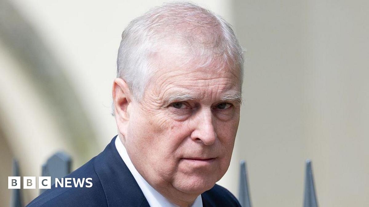 A close-up of Prince Andrew, who is outside in front of some railings - he is wearing a dark blue suit jacket and a white shirt which can just be seen and is looking at the camera with a neutral expression 