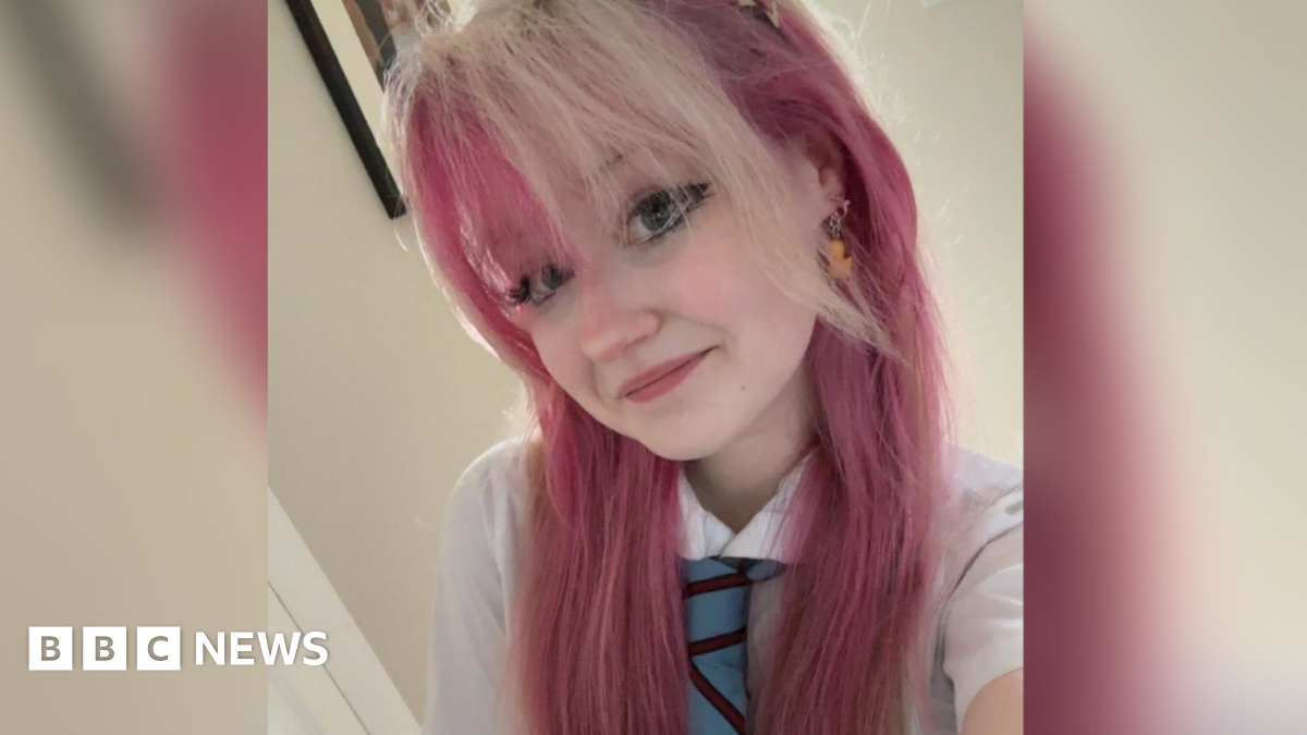 A selfie photograph of Holly with died pink hair wearing a white shirt and a blue and burgundy striped school tie. She has dark eye make up, rubber duck drop earrings and is smiling at the camera. 