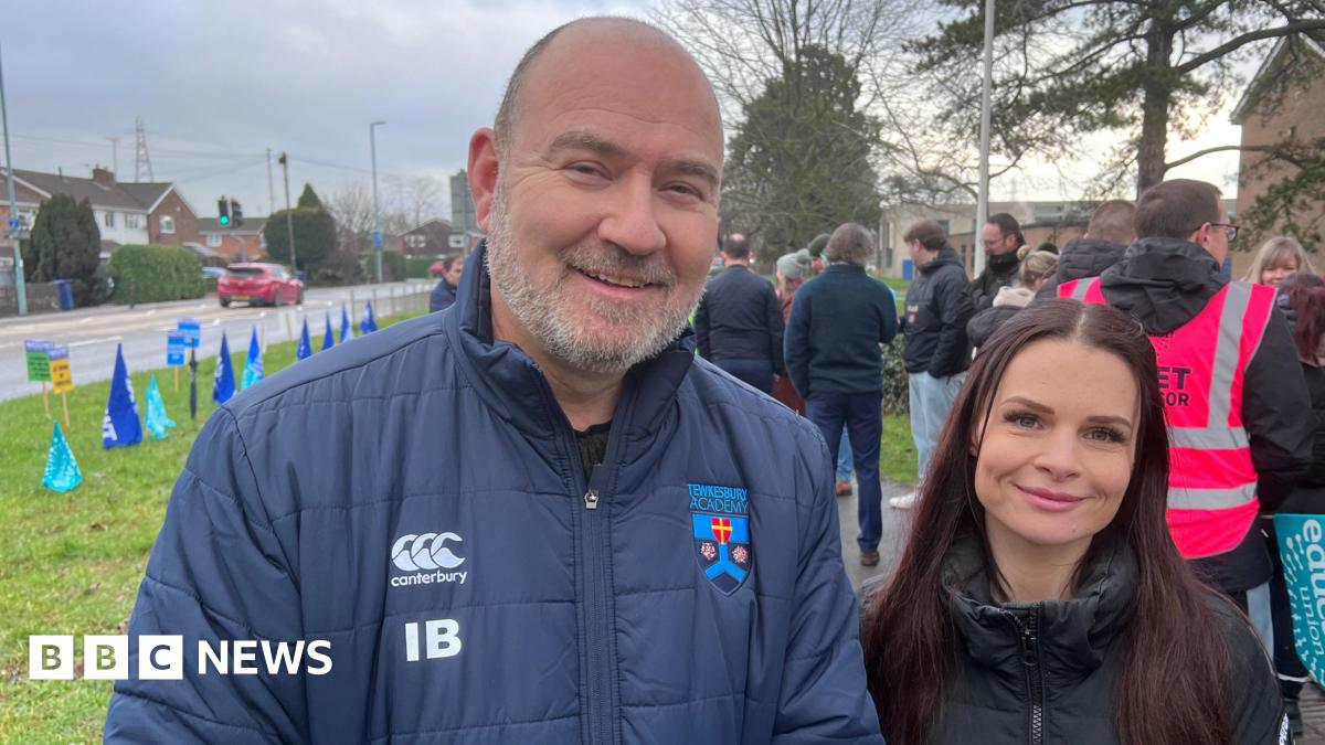A man with a small amount of short grey hair and a grey beard stands next to a woman with long, straight reddish brown hair. He is wearing a blue coat with the Tewkesbury Academy logo on it, along with others, and she is wearing a black coat.