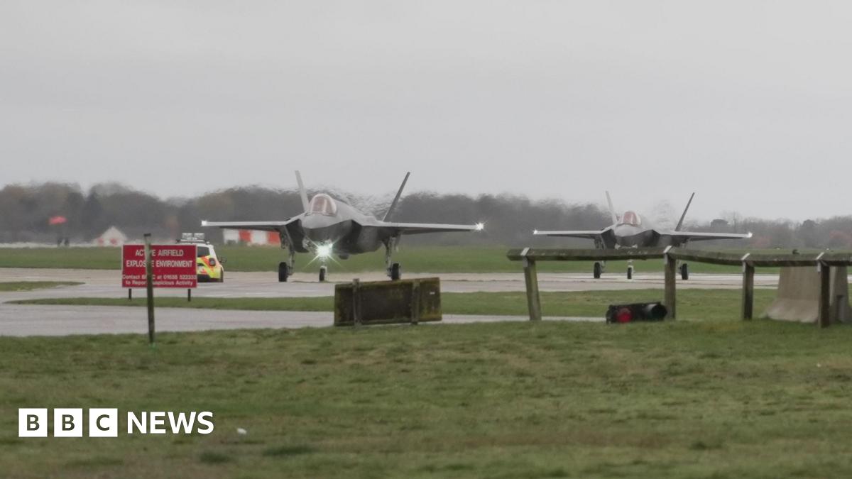 Two planes, which appear to be F-35As from the Joint Strike Fighter programme, located at RAF Lakenheath. They are located on a runway or taxiway, surrounded by grass lawns. A police car is visible. 