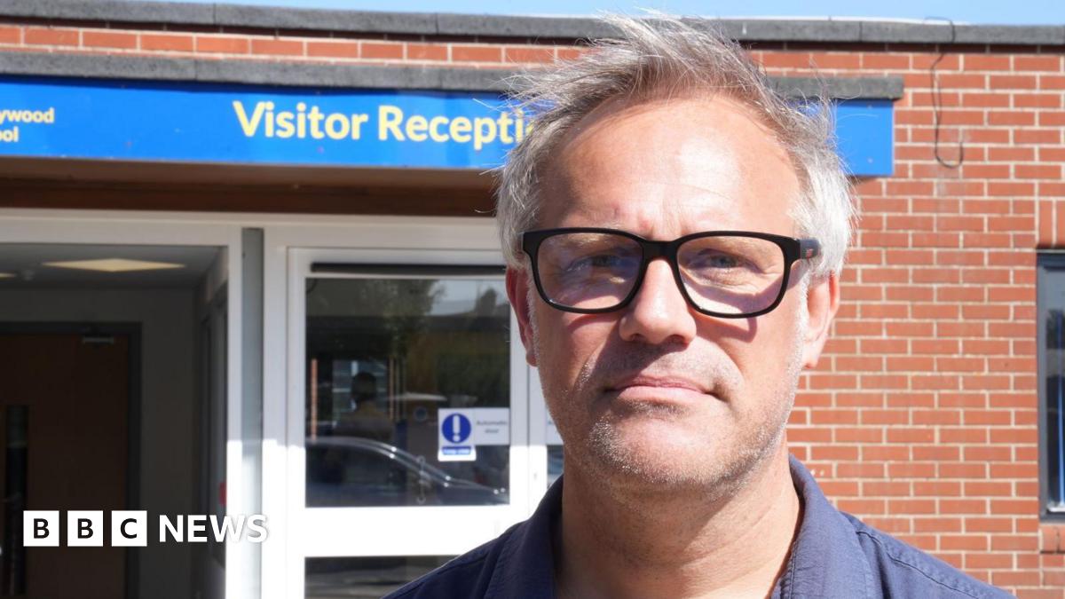 James Saunders, who has grey hair and is wearing glasses with black rims, standing outside the visitor entrance to Honywood School. The school is an orange brick building.