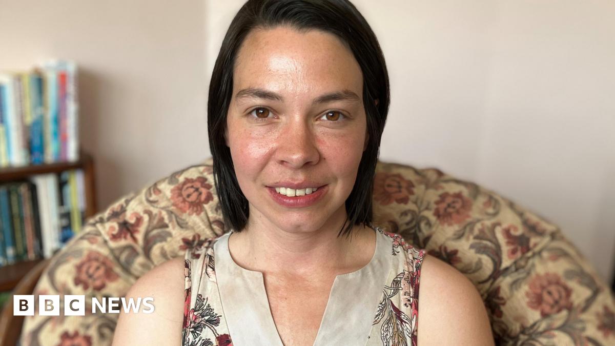 Stef Benstead with shoulder length brown hair and brown eyes is sitting on a chair with a floral pattern wearing a beige top and smiling. 
