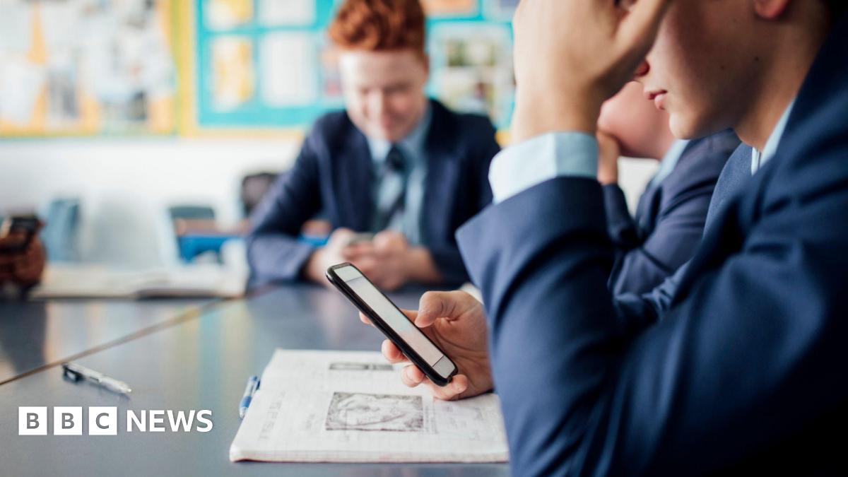 A child in a school uniform sat at a desk holding a smartphone.