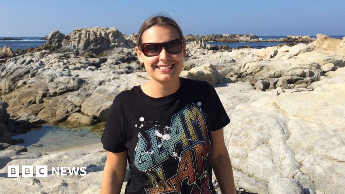 A woman is smiling, wearing sunglasses and a black graphic T-shirt, appears to be standing on some rocks at a seaside. 