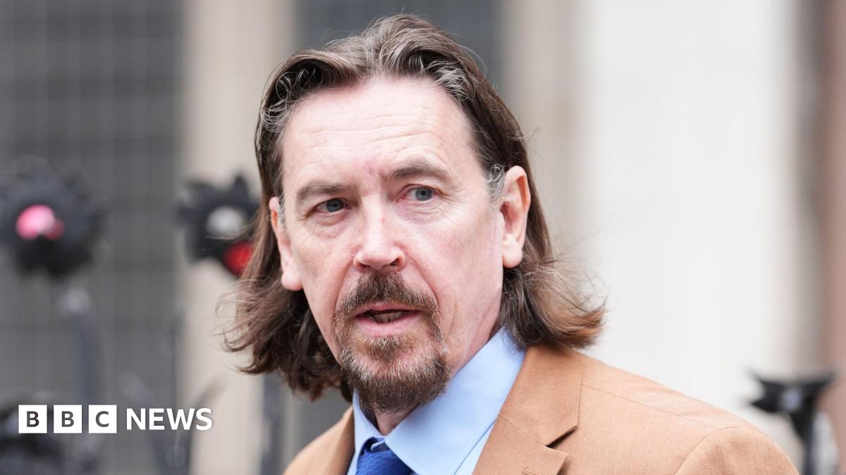Richard Hall wears a brown blazer and blue shirt and tie outside court. He has medium length brown hair and a brown moustache and beard.