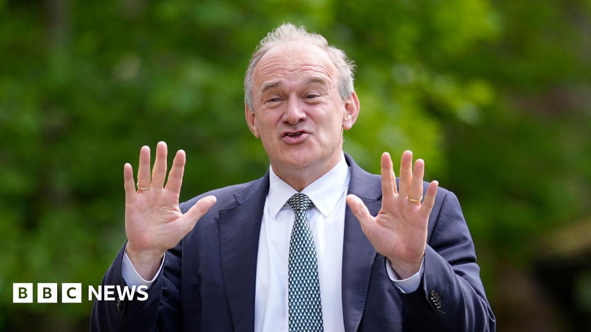 Ed Davey a middle aged man holding up his hands with a joyful expression. He is wearing a suit and tie. There is green foliage behind him.