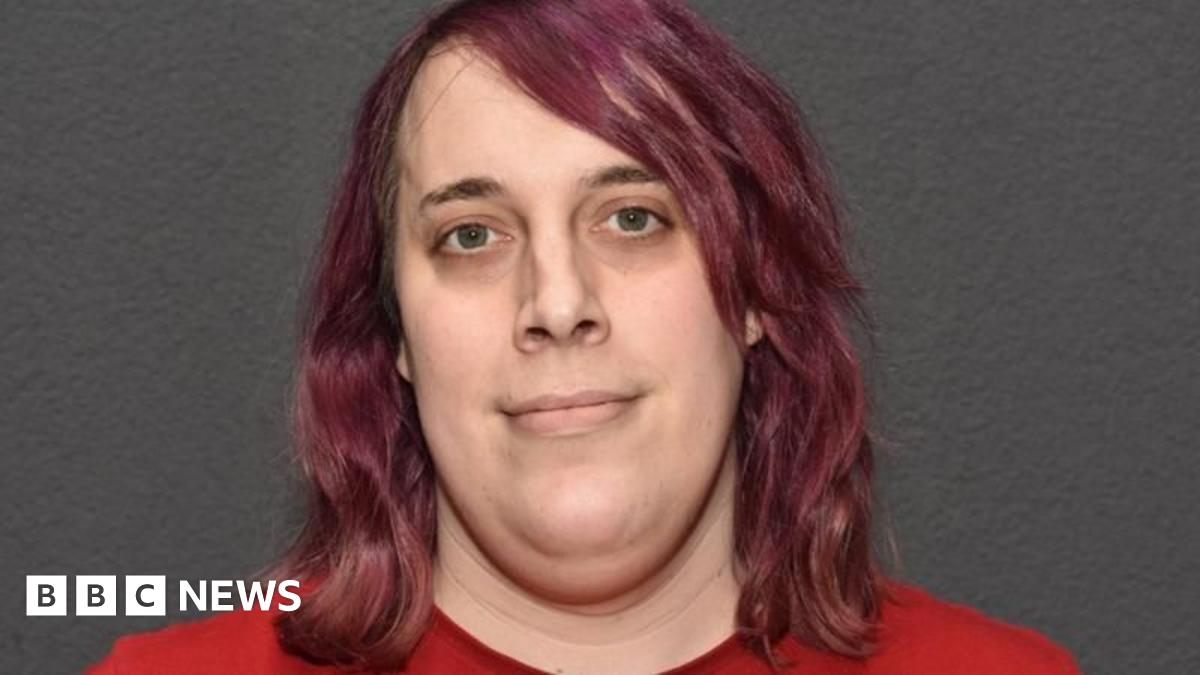 A police-issued image of Zoe Watts who has shoulder-length purple hair and is wearing a red top. She is standing in front of a grey wall.