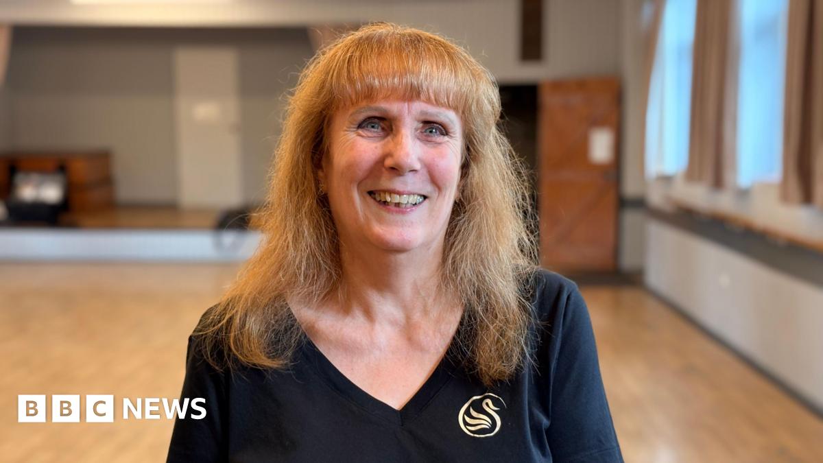 A medium close up of Glenys in a ballet studio. She is smiling wearing a black t-shirt with the polished wooden floor and a large mirror of the studio in soft focus in the background.