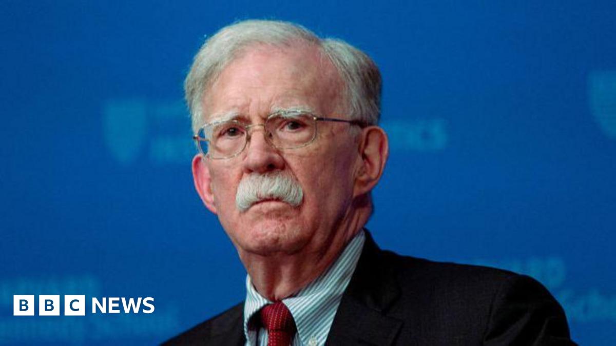A close-up image of John Bolton, who is looking straight ahead. He is wearing glasses, a black blazer, a stripped blue and white shirt and a red tie. 