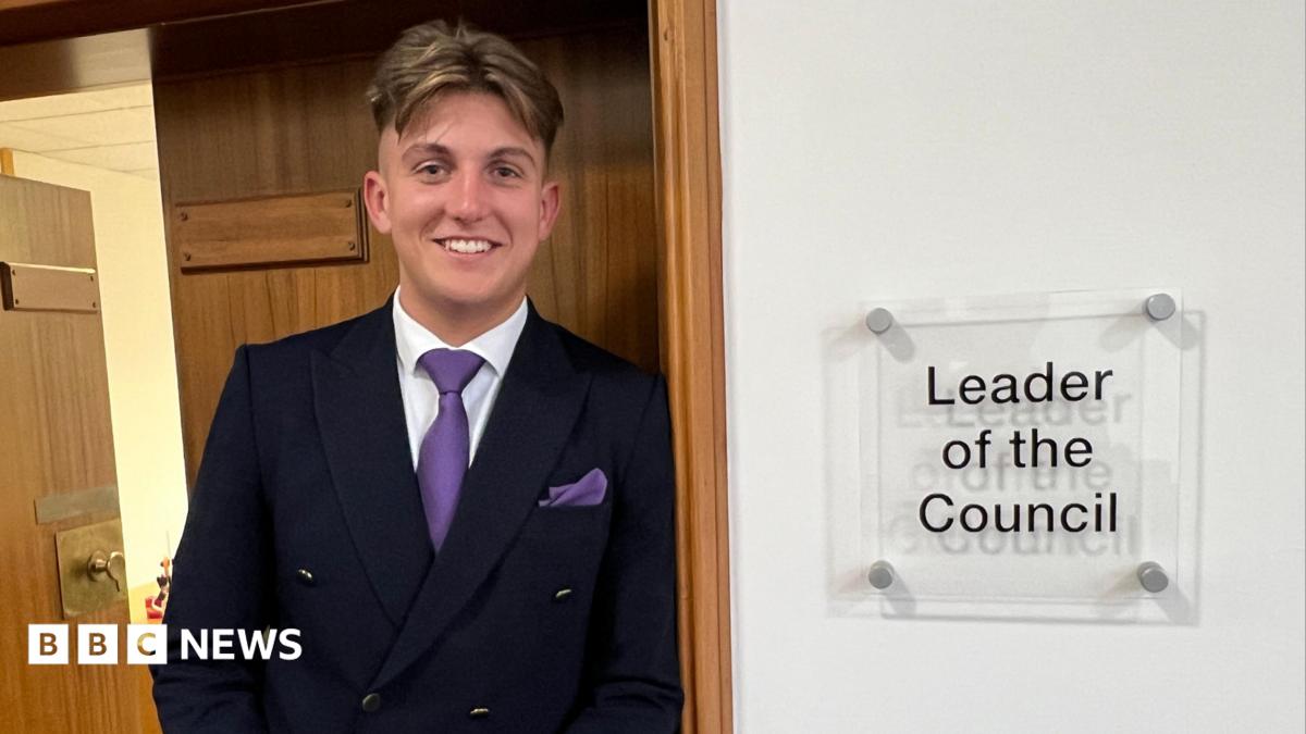 George Finch leans against a door frame. A sign to his left reads 'leader of the council'. He wears a dark suit, white shirt and purple tie.