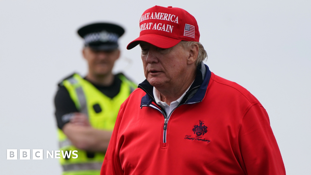 A man dressed in a red jumper and red hat, with the logo "make America great again" on it. In the background, a police officer stands with his arms folded. He is wearing a reflective vest and large black hat. 