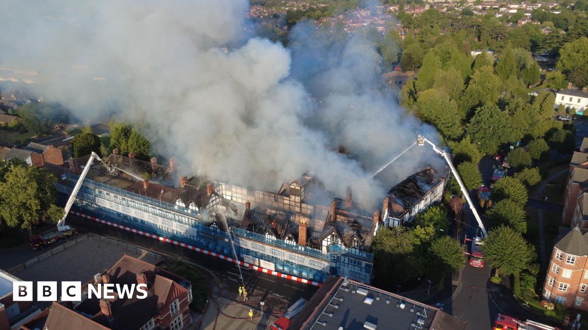 A black and white building engulfed in smoke. There are firefighters on two aerial platforms spraying water on it