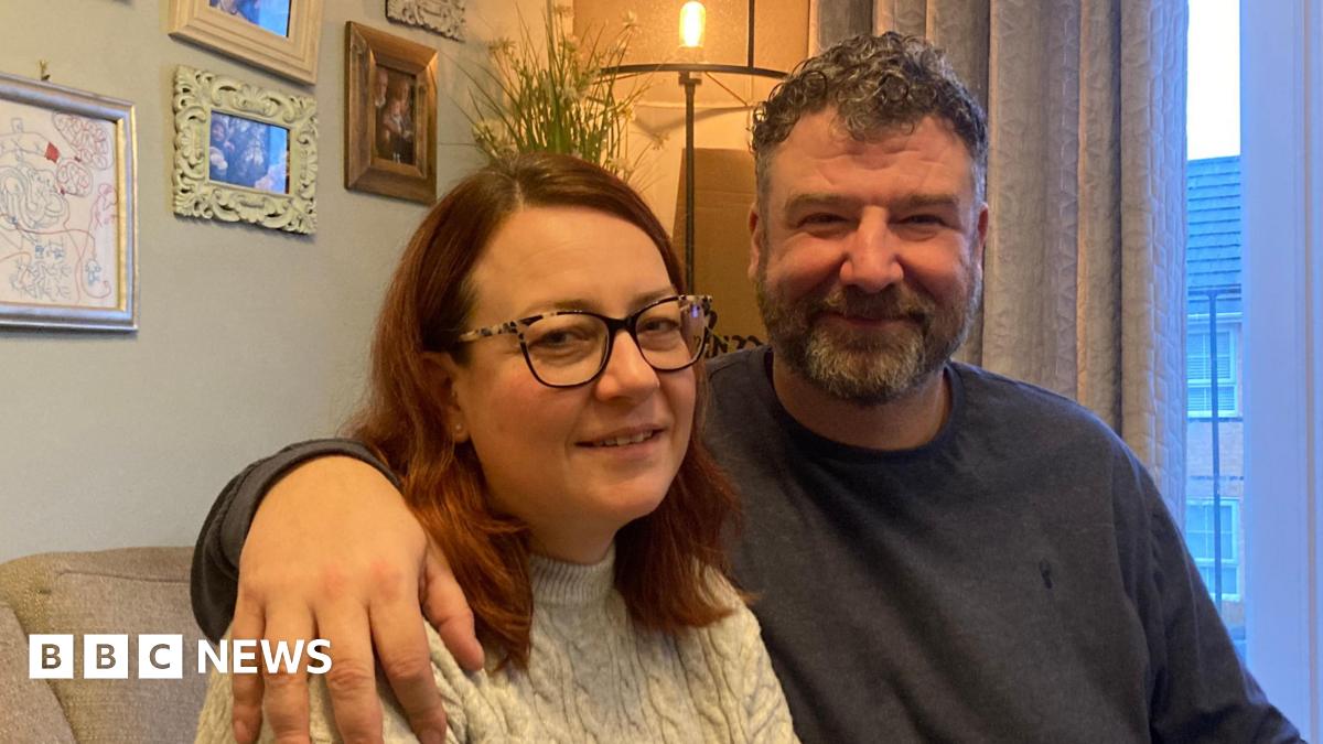 Kirsty and Donovan Shears sitting on a sofa, together at home. There are picture on the wall and they are both wearing jumpers and he has his arm around her shoulders.