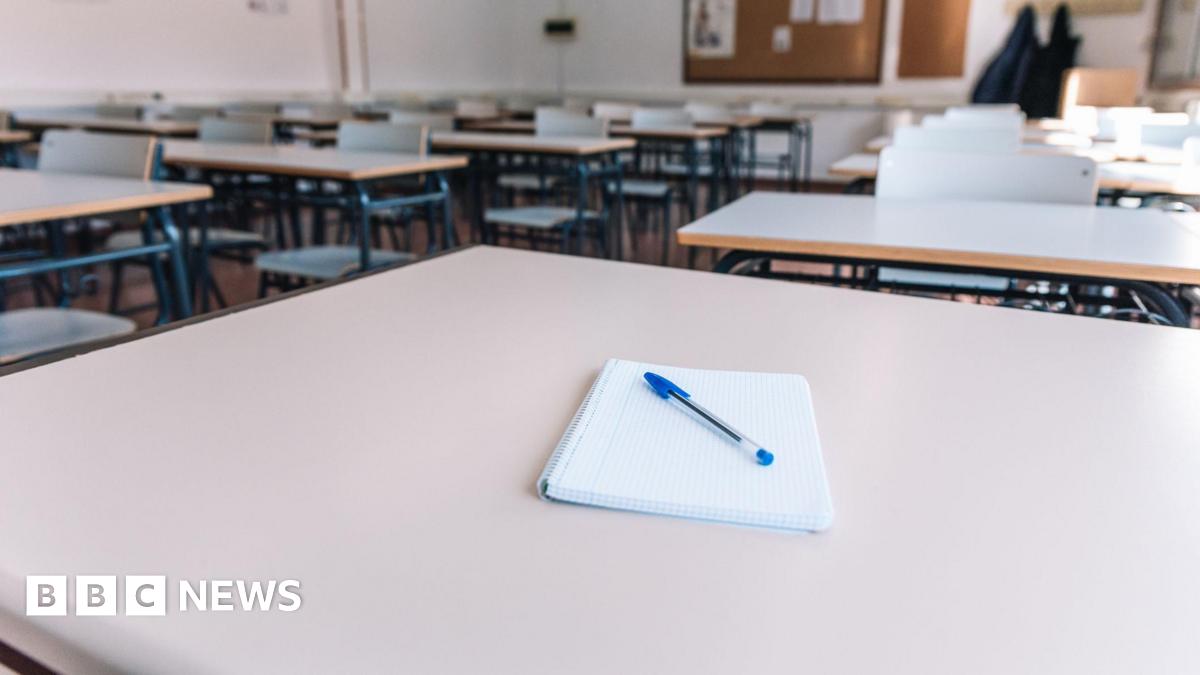 Stock image of a classroom. There are a number of grey desks. The one in the foreground has a notepad and pen on the top.