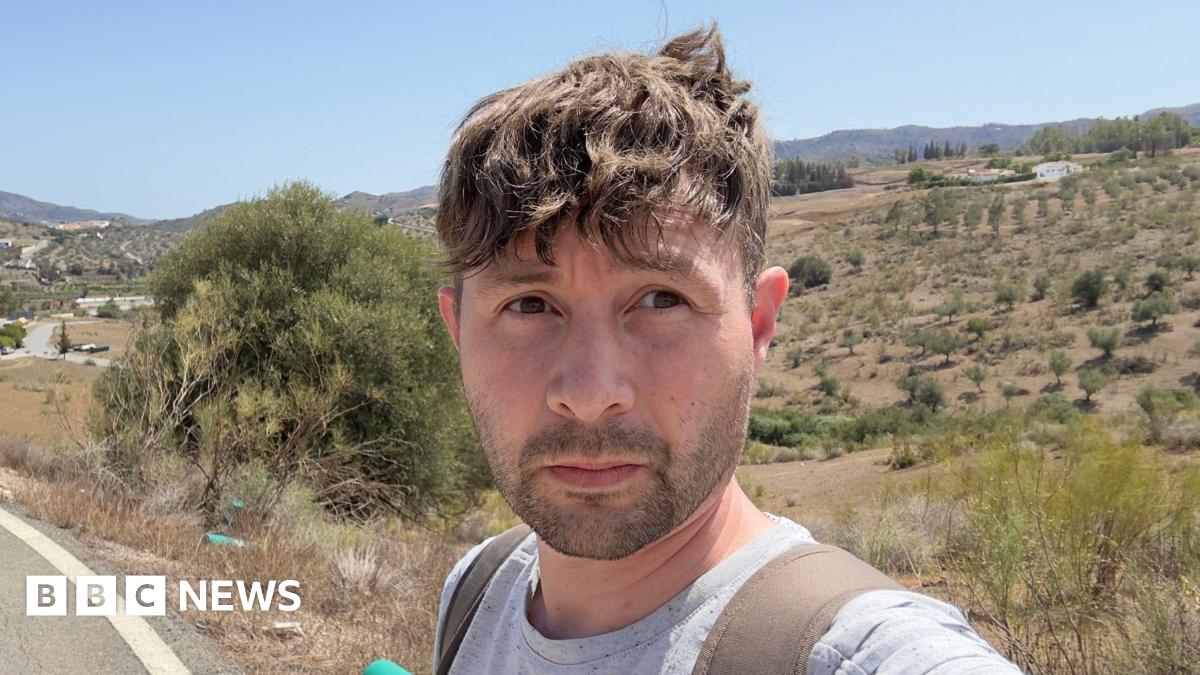 A selfie of a man with medium-length brown hair looking seriously at the camera against a backdrop of a Mediterranean landscape. You can see from his shoulders he's wearing a grey t-shirt and backpack.