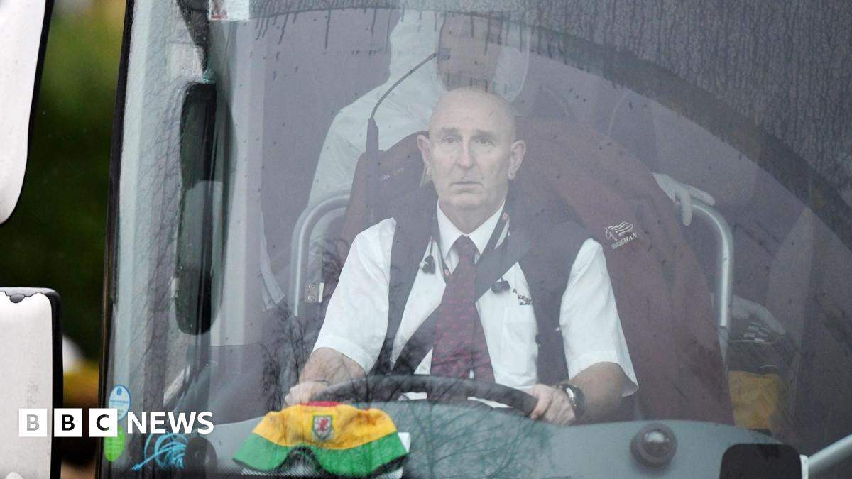 Andy Simonds sitting in the driving seat of the bus he drove, with a bucket Wales cap on the dashboard and people in PPE visible behind him