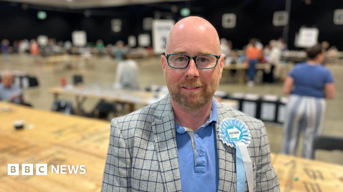 Nigel Clarke is wearing wearing glasses and a grey chequered jacket over a blue polo shirt. He is standing in the hall used for an election count