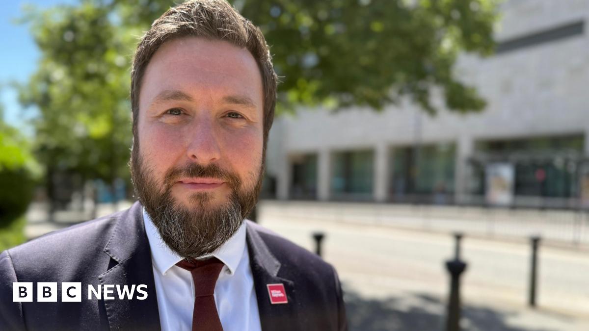 Pete Marland with short dark hair and beard, looking at the camera and wearing a black jacket, white shirt and dark tie. He is standing in front of trees and a road which passes a modern office building..